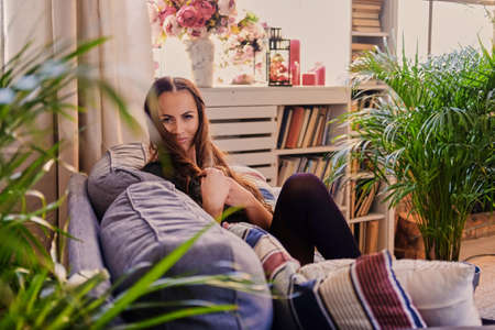 Portrait of a smiling brunette woman sits on a sofa in a room with green palm plants and book stands.の写真素材