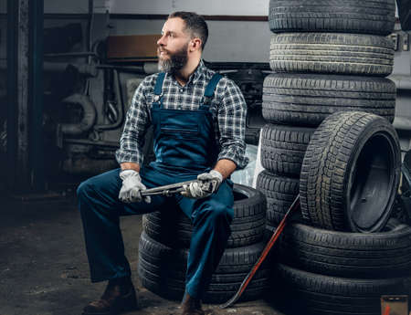 The brutal bearded mechanic male sits on an old tire in a garage.の写真素材