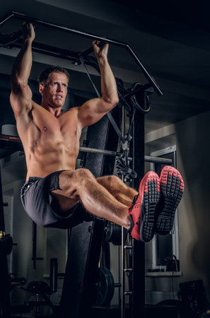Shirtless muscular man doing ABS workouts on pull up bar in a gym.
