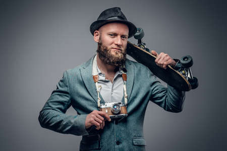 A stylish bearded hipster male dressed in a grey jacket and felt hat holds an SLR photo camera and a skateboard.の写真素材