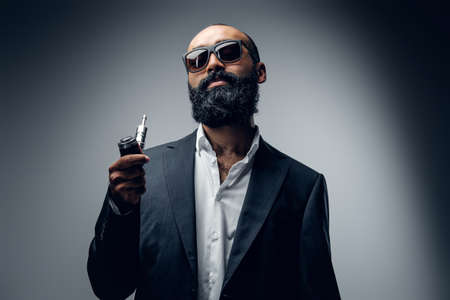 Hairless bearded male in sunglasses, dressed in a suit holds electronic cigarette.の写真素材