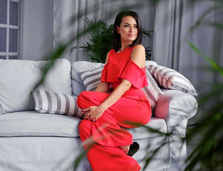 A brunette female dressed in a red evening dress sits on a coach in a room with green plants and grey interior.の写真素材
