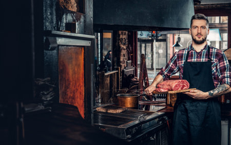 Portrait of a bearded chef with tattoos on his arms hold beef steak on a kitchen.の写真素材