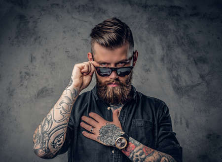 Studio portrait of a bearded hipster man with tattoos on his arms and neck  dressed in a black shirt and sunglasses