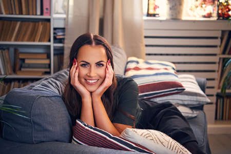 Portrait of positive female relaxing on a coach in a room with books stands and flowers.の写真素材