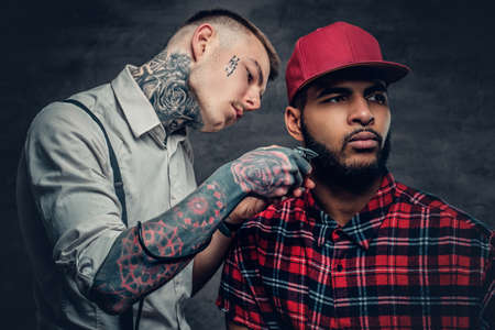 Tattooed barber cutting the beard of a Black hipster male in a baseball cap.の写真素材