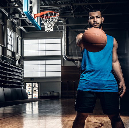 Portrait of Black basketball player holds a ball over a hoop in a basketball hall.の写真素材