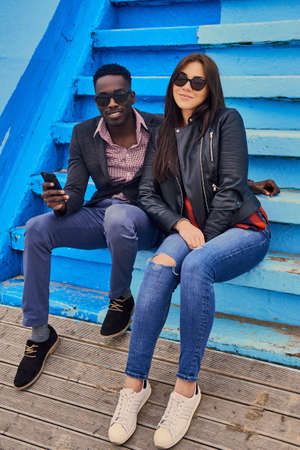 Stylish Black male and Caucasian female in sunglasses posing on a blue steps.