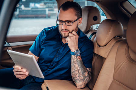 Stylish bearded male in eyeglasses with tattoo on his arm using portable tablet PC on a back seat of a car.の写真素材