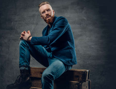 Portrait of redhead bearded hipster male dressed in a blue jacket posing on a wooden box over grey background.の写真素材