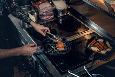A man with tattooed arm grilling a beef in a kitchen.の写真素材