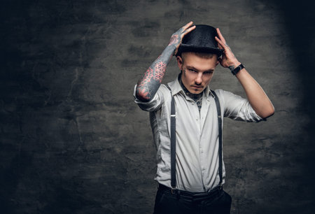 Stylish male with tattoos on his arms and face, dressed in a white shirt and top hat over grey background.の写真素材