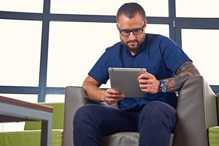 Bearded male in eyeglasses with tattoo on his arm sits on a chair and using a tablet PC.の写真素材