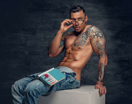 Portrait of shirtless muscular male with tattoo on a chest, dressed in a jeans and eyeglasses sits on a white wooden box and reads a book.の写真素材