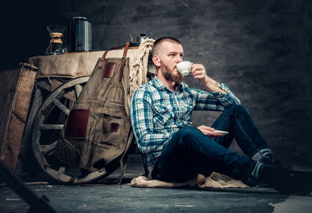 Redhead bearded hipster male dressed in a fleece shirt and jeans sits on a floor and drinks coffee.の写真素材