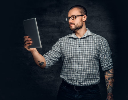 Bearded male in sunglasses holds tablet PC over grey background.