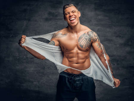 Studio portrait of smiling, athletic male with a tattoo on his chest ripping t shirt.の写真素材