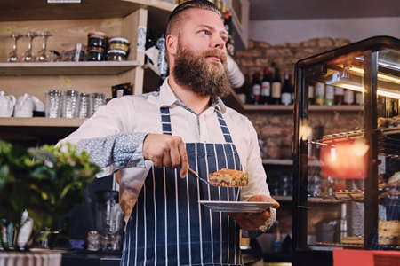 The stylish bearded male holds chocolate cake at the counter in the bakery shop.の写真素材