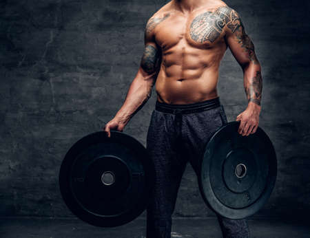 The muscular shirtless, tattooed male holds barbell weights over grey background.の写真素材
