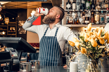 The bearded coffee seller holds a coffee cup at the counter in a coffee shop.