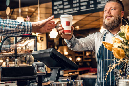 Positive bearded barista male selling coffee to a consumer in a coffee shop.の写真素材