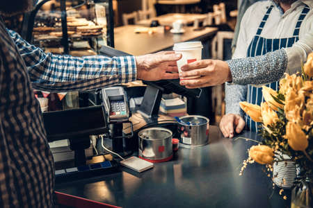 Positive bearded barista male selling coffee to a consumer in a coffee shop.の写真素材