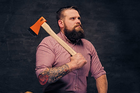 Bearded lumberjack male with tattoos on arms, sits on a wooden box and holds the axe.