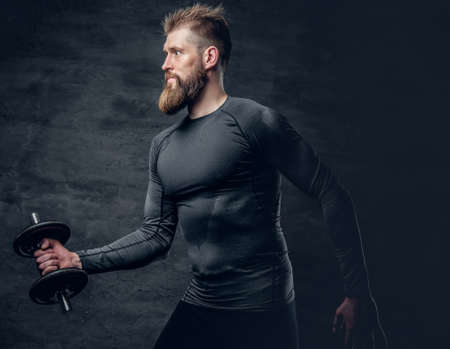 Studio portrait of sporty bearded male dressed in a grey sportswear holds dumbbell.の写真素材