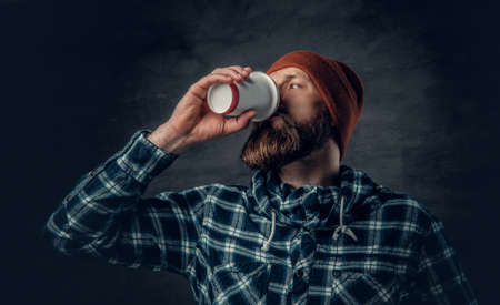 A brutal bearded male dressed in a hat and fleece shirt, drinks coffee from a paper glass.の写真素材