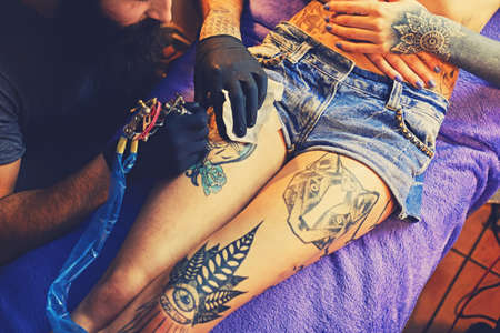 The bearded tattoo artist makes a tattoo on a woman's leg in an ink saloon.の写真素材