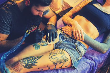 The bearded tattoo artist makes a tattoo on a woman's leg in an ink saloon.の写真素材