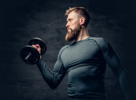 Studio portrait of sporty bearded male dressed in a grey sportswear holds dumbbell.の写真素材