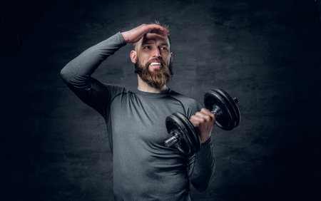 Studio portrait of sporty bearded male dressed in a grey sportswear holds dumbbell.