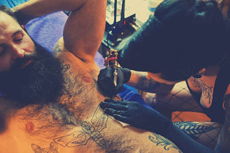 A professional female tattoo artist makes black and white tattoo on bearded male torso in a salon.の写真素材