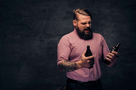 A bearded fat male dressed in a pink fleece shirt holds a beer bottle over dark grey background.の写真素材