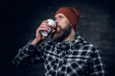 A brutal bearded male dressed in a hat and fleece shirt, drinks coffee from a paper glass.の写真素材