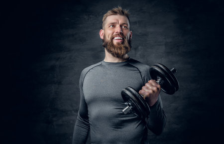 Studio portrait of sporty bearded male dressed in a grey sportswear holds dumbbell.の写真素材