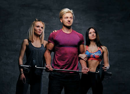 Threesome fitness model of blond and brunette slim women and a man holds barbell posing over grey background.の写真素材