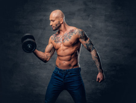 Positive shaved head shirtless male dressed in a jeans holds the dumbbell over dark grey background.の写真素材