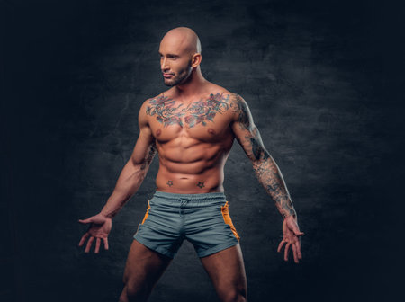 Shaved head shirtless muscular male with tattooed body dressed in a sport short over grey background.の写真素材