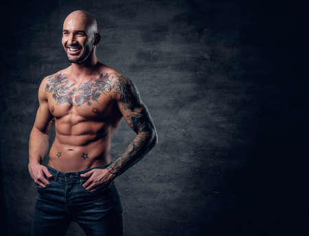 Studio portrait of smiling shirtless shaved head, muscular male with tattoos on his torso over grey vignette background.の写真素材
