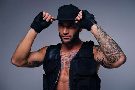 Tattooed male dressed in a jeans, cap and waistcoat on a naked torso isolated on a grey vignette background.の写真素材