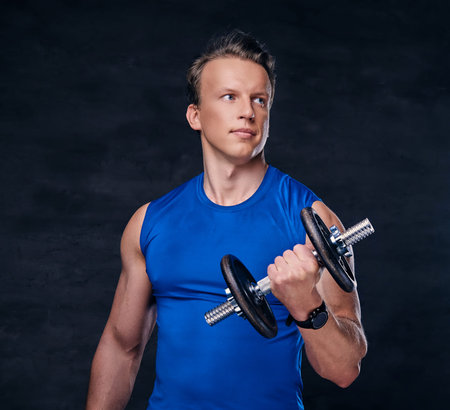 Portrait of athletic male dressed in a blue sportswear holds the dumbbell over dark grey background.