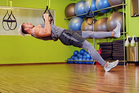 Athletic muscular blond male exercising with trx straps in a gym club.
