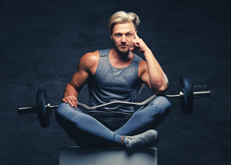 A blond, athletic male dressed in a grey sportswear sits on a white wooden box and holds barbel.の写真素材