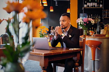 A thoughtful black American male dressed in an elegant suit using a laptop in a cafe.