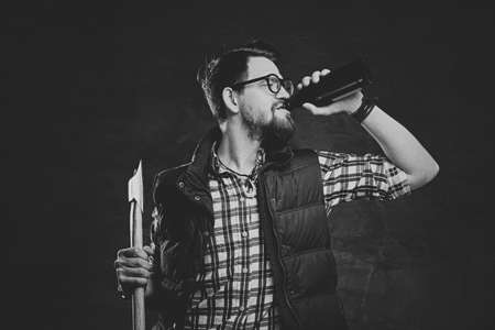 Portrait of funky bearded hipster male dressed in fleece shirt holds an axe and drinks craft bottled beer.の写真素材