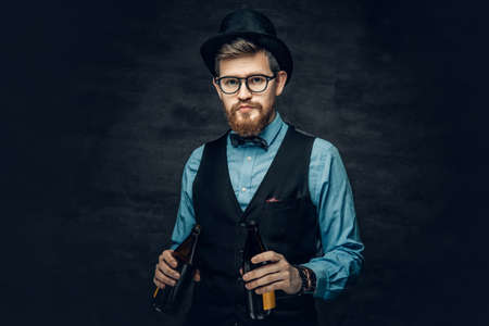Portrait of bearded hipster male dressed in a blue shirt, elegant waistcoat and top hat holds two craft beer bottles with a think of a party.の写真素材