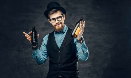 Portrait of bearded hipster male dressed in a blue shirt, elegant waistcoat and top hat holds two craft beer bottles with a think of a party.の写真素材