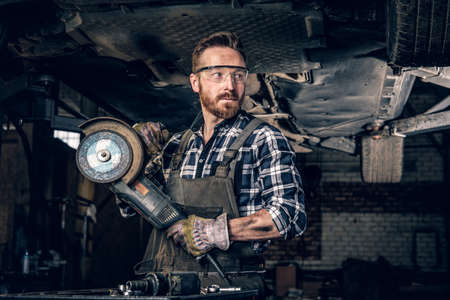 Bearded mechanic in protective googles holds angle grinder near the car in a workshop.の写真素材